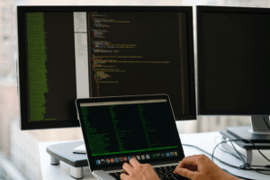Developer coding at a desk with multi-monitor setup, terminal output and code visible, modern office background